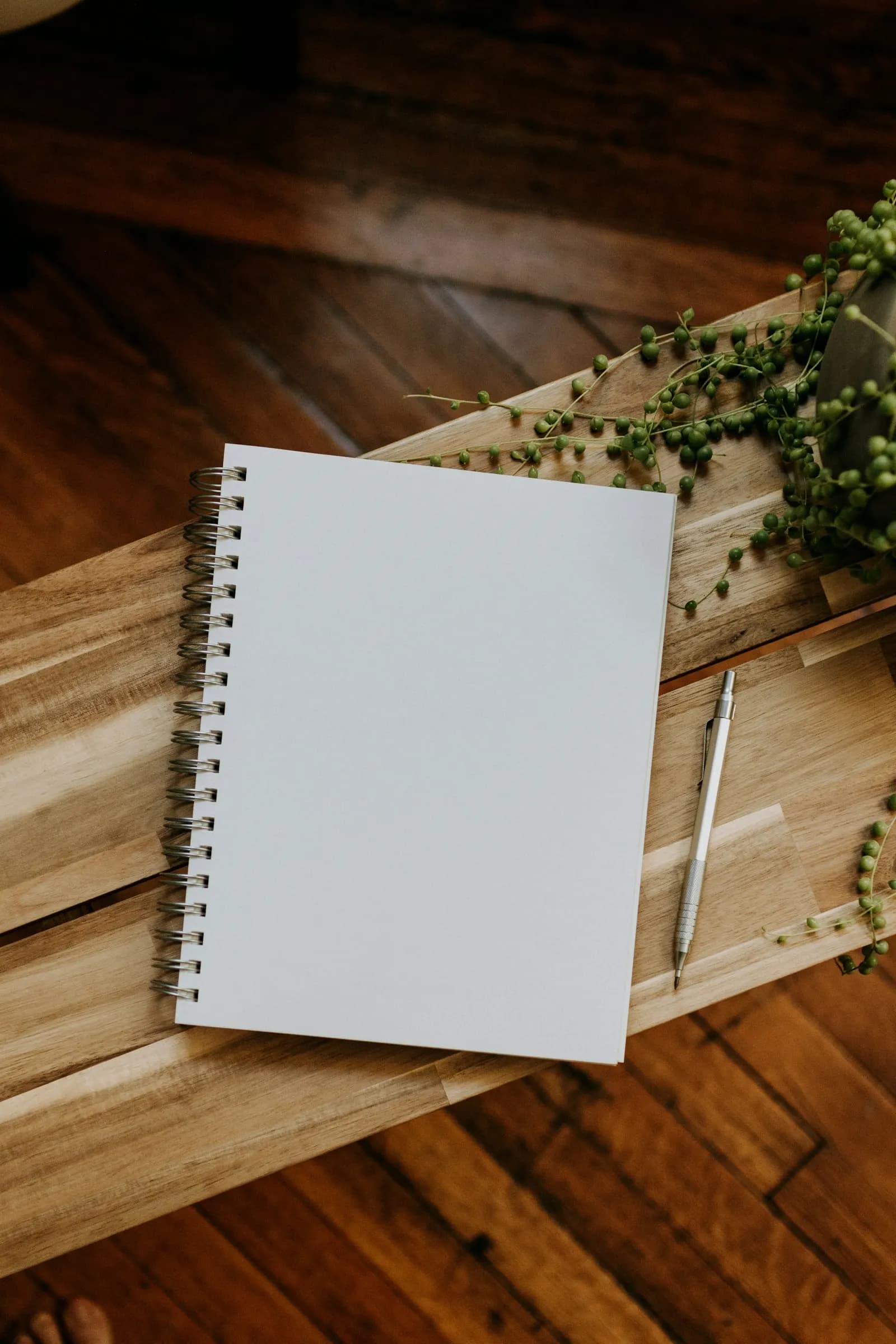 A white spiral notebook and a pencil lying on a wooden board over a wooden floor.