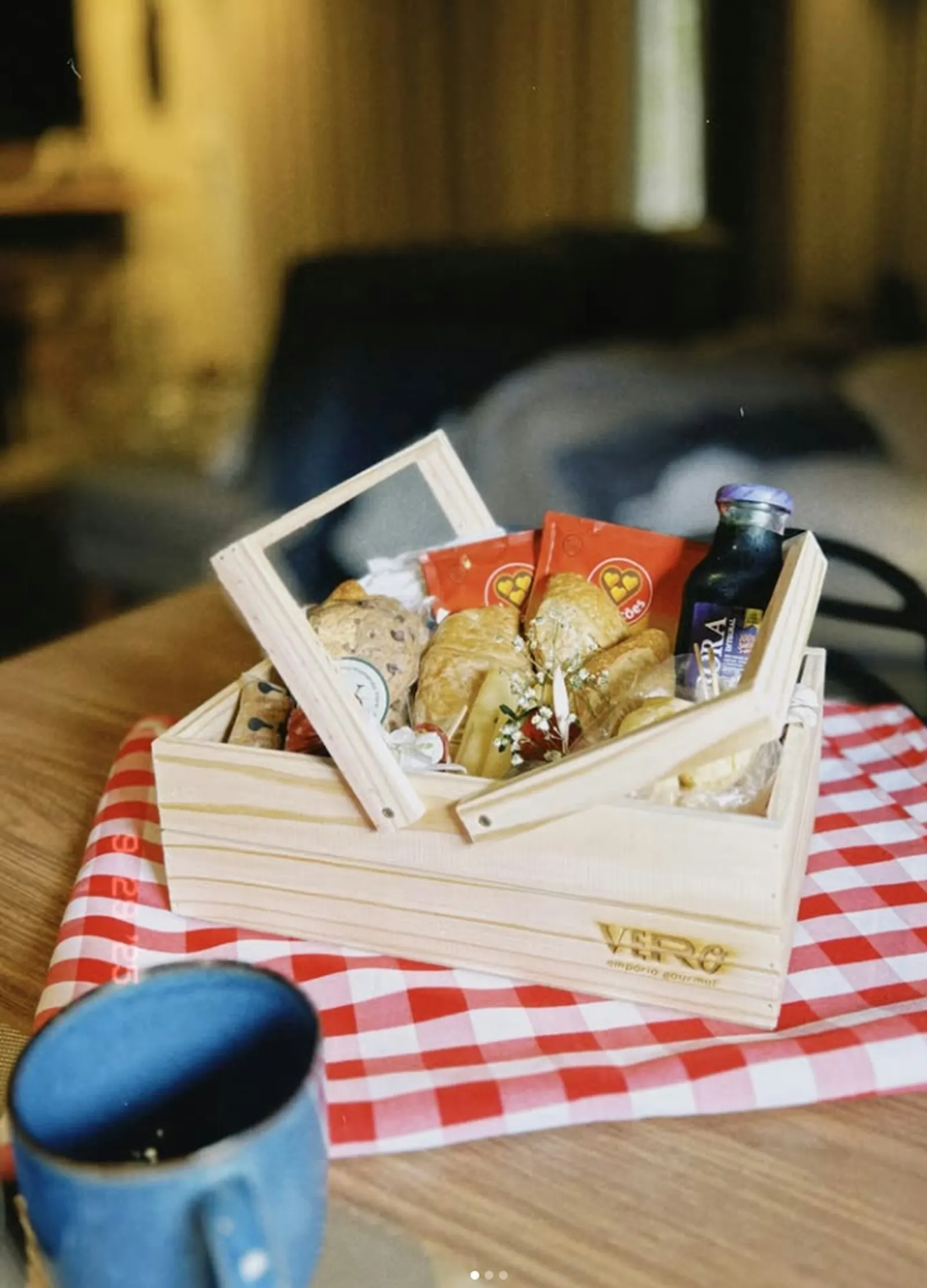 A wooden basket with a clear lid, filled with pastries and a small bottle of juice, on a red checked cloth.