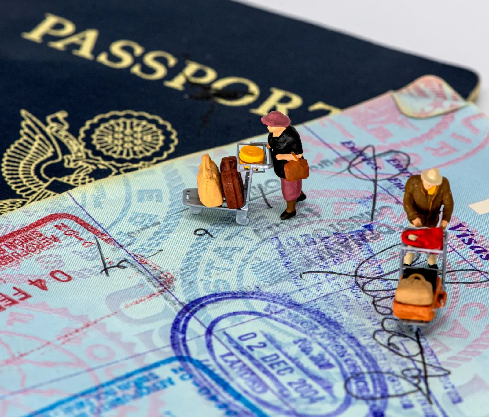 Two tiny figures with luggage carts standing on an open passport page covered in travel stamps.