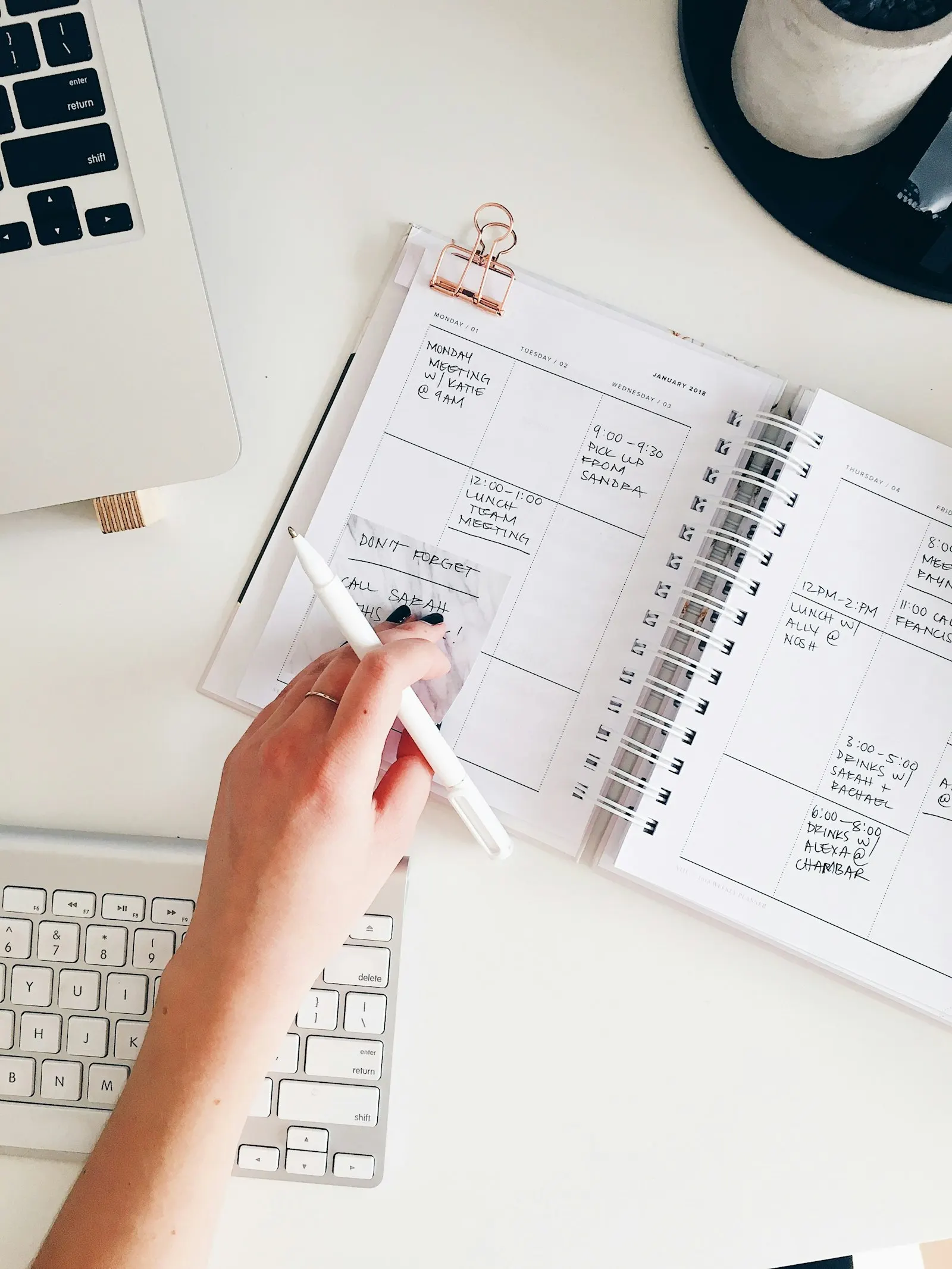 A person writing in a spiral planner on a white desk next to a laptop.