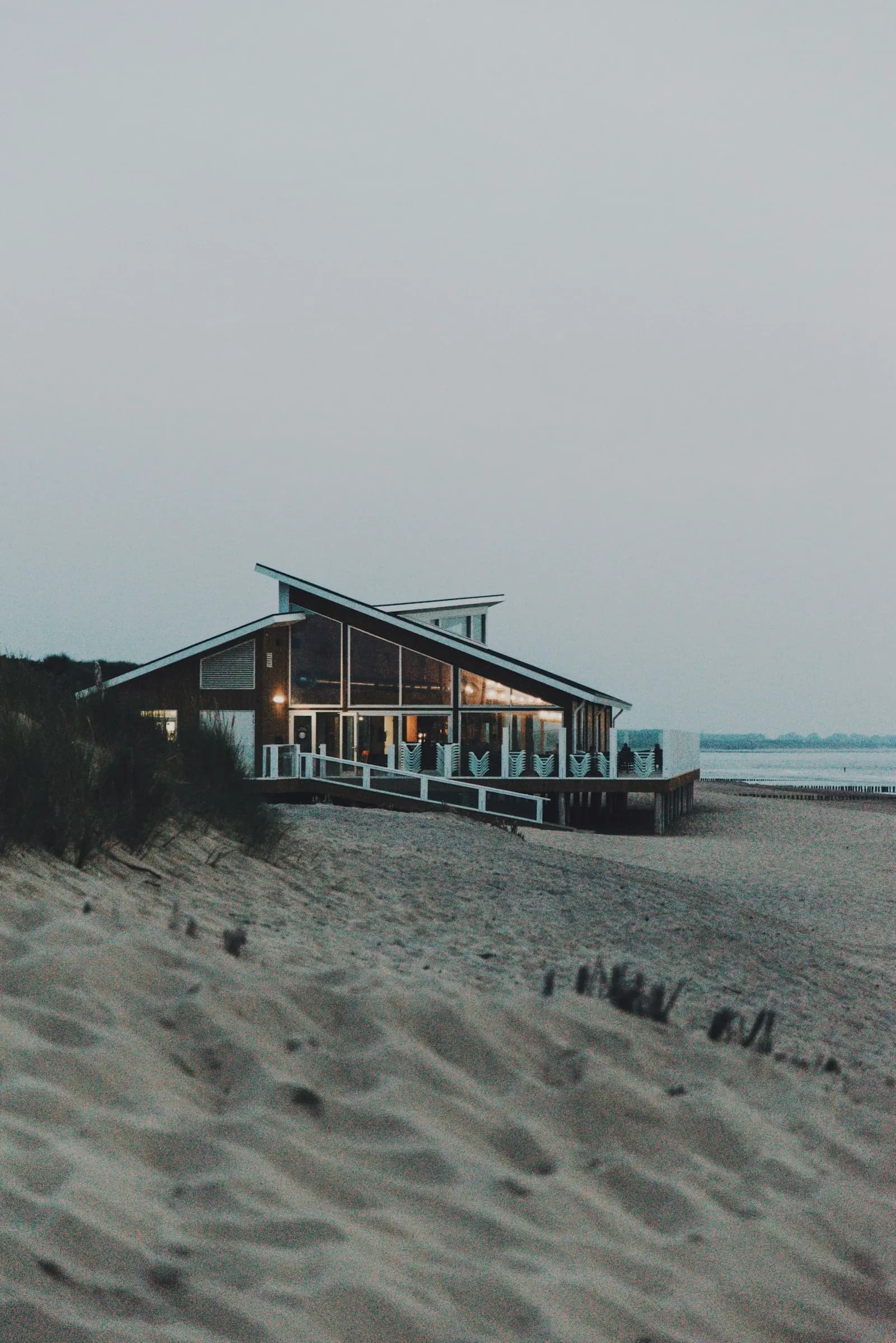 A modern holiday house on a sandy beach at dusk, with warm light in the windows.