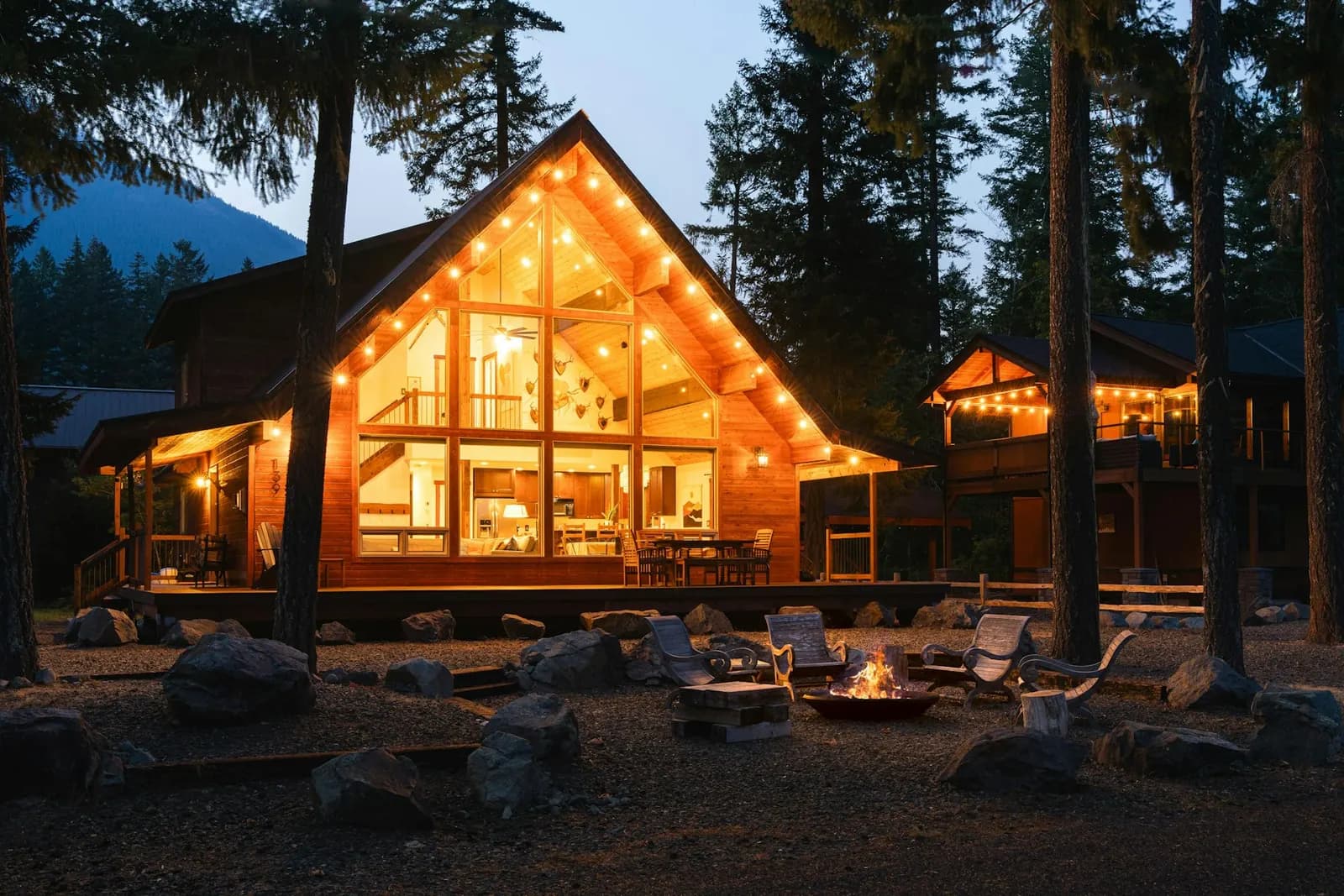 A glowing wooden cabin with large windows at dusk, with a fire pit in front.